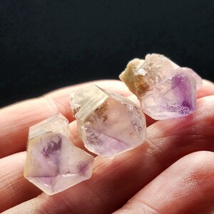 Puede incluir: Tres cristales de amatista en bruto, con tonos morados, beige y transparentes. Los cristales tienen una textura natural y rugosa, sobre un fondo oscuro.