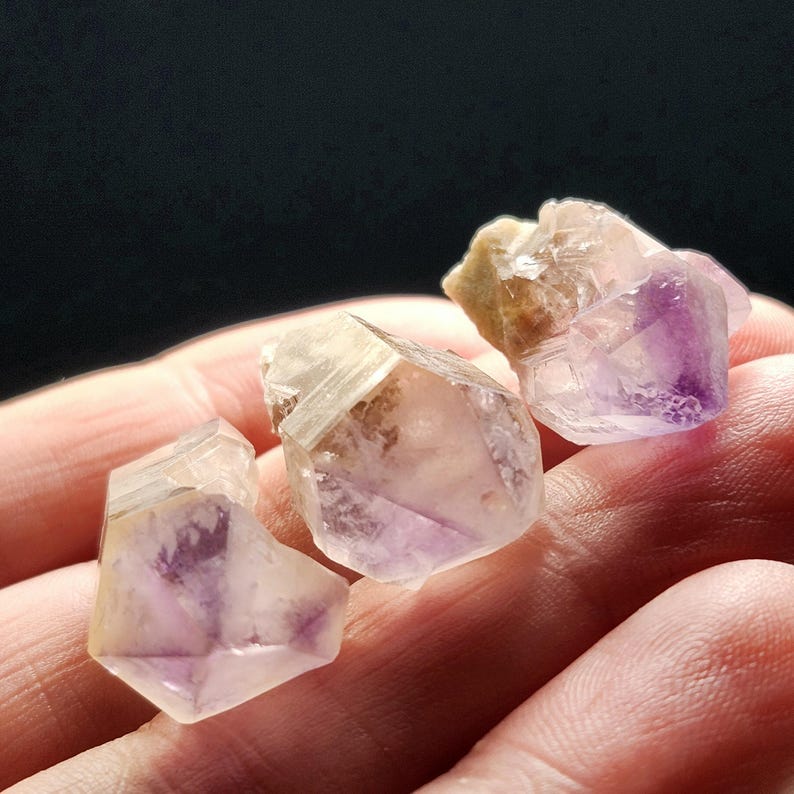 Puede incluir: Tres cristales de amatista en bruto, con tonos morados, beige y transparentes. Los cristales tienen una textura natural y rugosa, sobre un fondo oscuro.