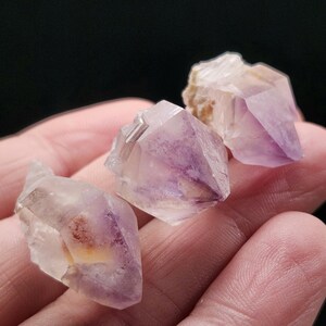 Puede incluir: Tres grupos de cristales de amatista, que muestran tonos p&uacute;rpuras y blancos transl&uacute;cidos. Los cristales tienen una textura natural y rugosa. La imagen muestra los cristales sobre un fondo oscuro.