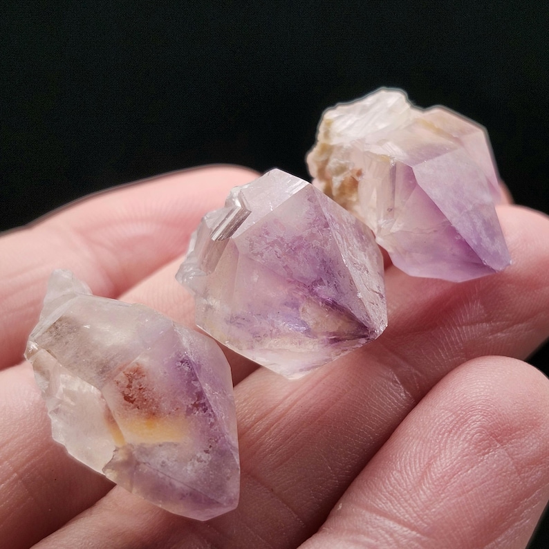 Puede incluir: Tres grupos de cristales de amatista, que muestran tonos p&uacute;rpuras y blancos transl&uacute;cidos. Los cristales tienen una textura natural y rugosa. La imagen muestra los cristales sobre un fondo oscuro.