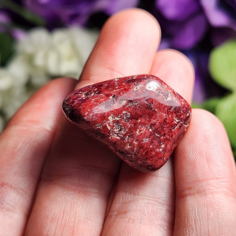 May include: A red, irregularly shaped tumbled stone with a smooth, polished surface. The stone has a speckled appearance with a mix of darker and lighter red hues.
