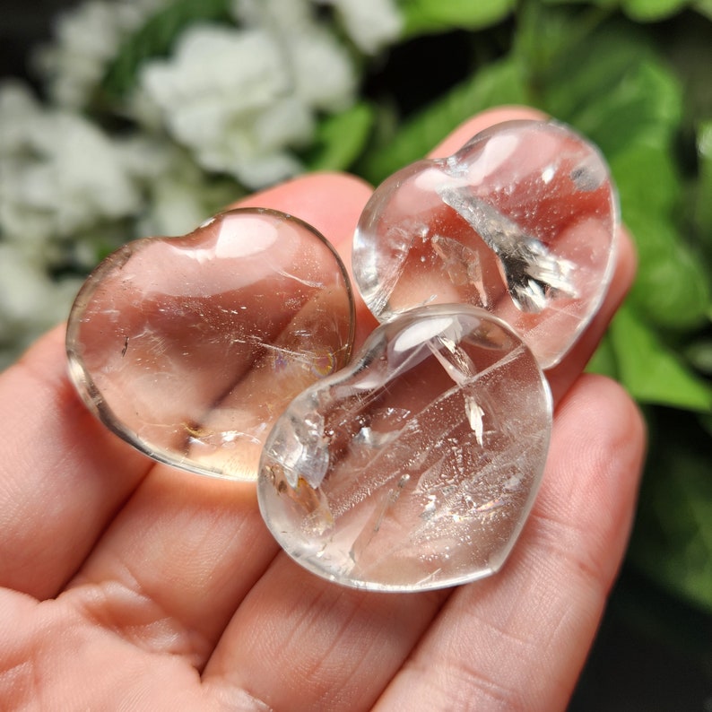May include: Three clear quartz crystal hearts with a slight golden hue. The hearts are smooth and polished, and they are arranged in a hand.