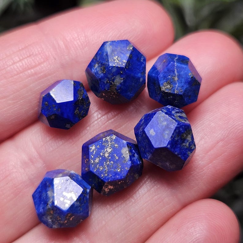May include: Six faceted lapis lazuli gemstones with a deep blue color and natural gold-colored pyrite inclusions. Each stone features a multi-faceted geometric cut, displaying a polished surface that reflects light across the various angled faces.