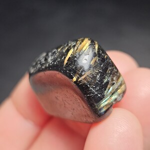 May include: A polished, black gemstone with gold and iridescent inclusions. The stone has a smooth, rounded shape with a textured surface on one side. The gemstone is held in a person's hand.