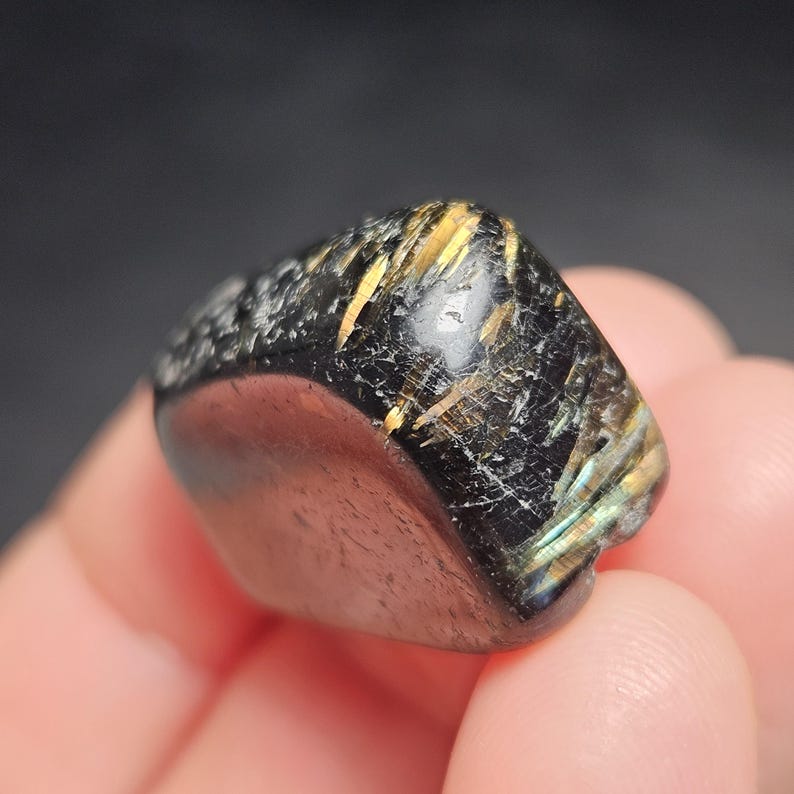 May include: A polished, black gemstone with gold and iridescent inclusions. The stone has a smooth, rounded shape with a textured surface on one side. The gemstone is held in a person's hand.