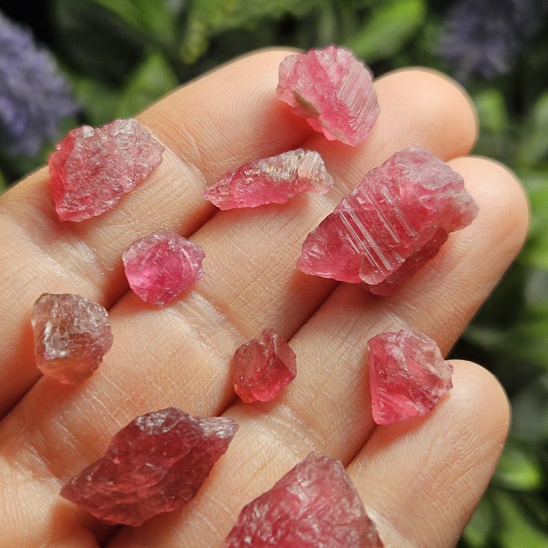 May include: A collection of small, raw, unpolished pinkish-red tourmaline crystals with irregular shapes and faceted surfaces. These translucent to opaque mineral specimens display varying shades of deep pink and magenta with natural textures.