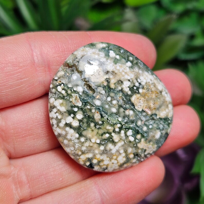 May include: A polished, oval-shaped gemstone with a mottled pattern of green, white, and tan. The stone has a smooth surface and a natural, organic appearance. The colors are distributed in a speckled and swirled design.