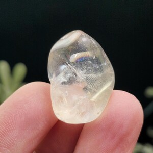 May include: A clear, polished gemstone with a rounded shape. The stone has a pale yellow hue and internal feather-like inclusions. The gemstone is held between two fingers, with a dark background.