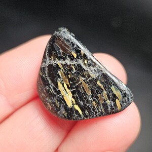 May include: A polished, triangular gemstone with a black base color and striking gold and white veining. The stone is held in a hand, showcasing its natural beauty and unique pattern. The gemstone is a natural mineral.