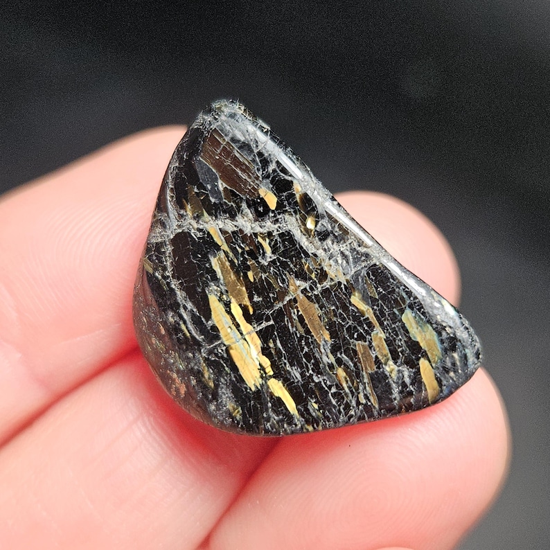 May include: A polished, triangular gemstone with a black base color and striking gold and white veining. The stone is held in a hand, showcasing its natural beauty and unique pattern. The gemstone is a natural mineral.