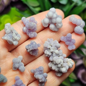 May include: A selection of small, grape-like clusters of pale purple chalcedony gemstones. The stones have a smooth, rounded texture and vary slightly in size and shape. The background is a blurred mix of green and purple.