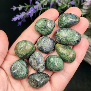 May include: A collection of polished green and blue stones. The stones have a smooth, rounded shape and a marbled appearance, with varying shades of green and blue. The stones are held in a hand, with purple flowers in the background. The stones are approximately 1-2 inches in size.