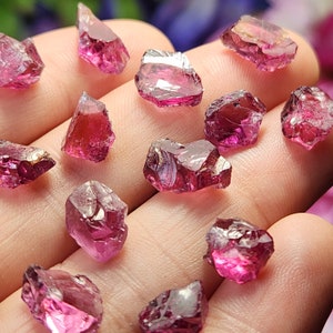 May include: A collection of pink and red rough gemstones. The gemstones are various shapes and sizes and have a natural, unpolished look.