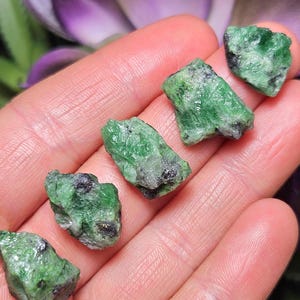 May include: A collection of raw emerald gemstones, each a vivid green with dark inclusions. The gemstones are held in a hand, with a blurred purple flower in the background. The emeralds are of varying sizes and shapes, showcasing their natural form.