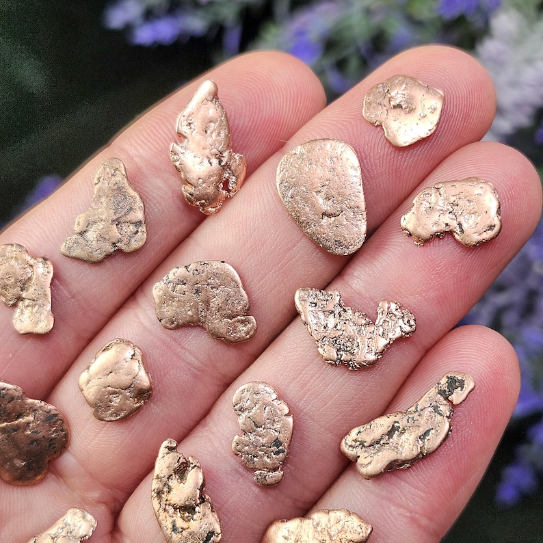 May include: A collection of small, irregularly shaped gold nuggets. The nuggets vary in size and texture, with a warm, metallic gold color. The image is taken in natural light, highlighting the details of the gold.