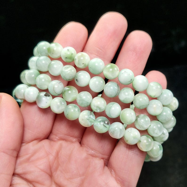 Peut inclure: Trois rang&eacute;es de bracelets de perles vert clair. Les perles rondes ont un aspect marbr&eacute; avec des variations de couleur. Les bracelets sont tenus dans une main sur fond noir. Les perles mesurent environ 0,8 cm de diam&egrave;tre.