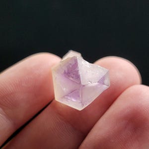 Puede incluir: Primer plano de un peque&ntilde;o cristal facetado con un exterior amarillo p&aacute;lido y un interior p&uacute;rpura. El cristal se sostiene entre dos dedos sobre un fondo oscuro. La forma geom&eacute;trica del cristal es claramente visible.