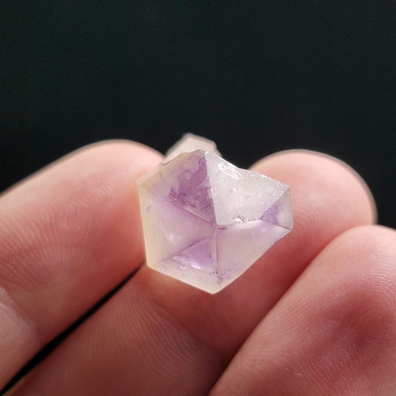 Puede incluir: Primer plano de un peque&ntilde;o cristal facetado con un exterior amarillo p&aacute;lido y un interior p&uacute;rpura. El cristal se sostiene entre dos dedos sobre un fondo oscuro. La forma geom&eacute;trica del cristal es claramente visible.