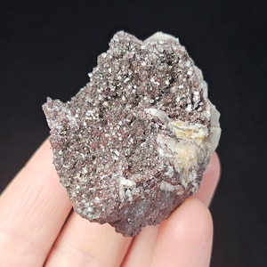 May include: A natural mineral specimen featuring a dark reddish-brown matrix with sparkling, silvery-white crystal clusters. The mineral has a rough, textured surface and is held in a hand against a black background.