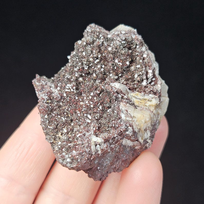 May include: A natural mineral specimen featuring a dark reddish-brown matrix with sparkling, silvery-white crystal clusters. The mineral has a rough, textured surface and is held in a hand against a black background.