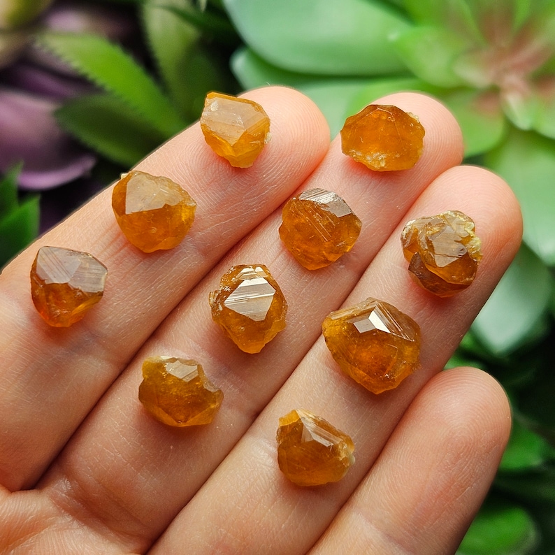 May include: A collection of raw, golden-orange gemstones with faceted surfaces. The crystals vary in size and shape, displaying a natural, unpolished appearance. The gemstones are arranged on a light-colored surface, with a blurred green background.