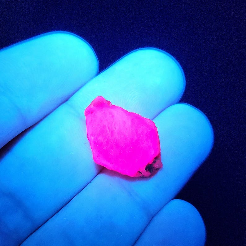 May include: A bright pink, irregularly shaped, rough-cut gemstone under a UV light.