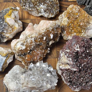 May include: A collection of various raw mineral specimens. The stones display a range of colors, including white, brown, yellow, and dark red. Some have crystalline structures, while others have a rough, textured appearance. The minerals are arranged on a brown surface.