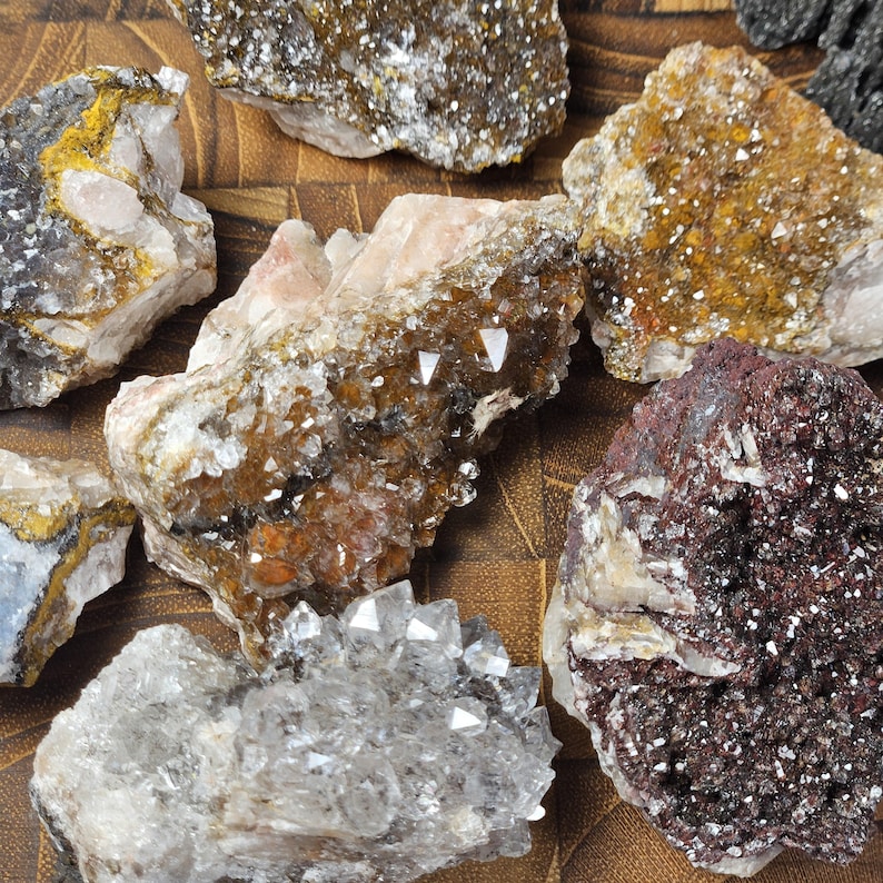 May include: A collection of various raw mineral specimens. The stones display a range of colors, including white, brown, yellow, and dark red. Some have crystalline structures, while others have a rough, textured appearance. The minerals are arranged on a brown surface.