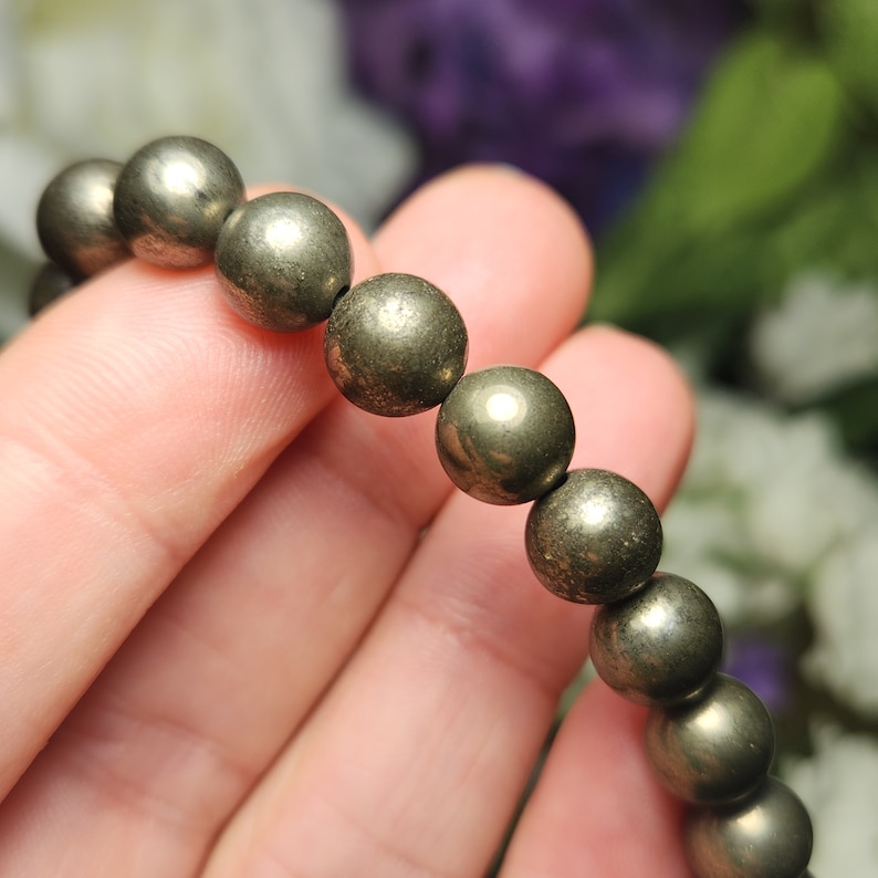 May include: A pyrite bracelet with spherical, metallic beads. The beads have a textured, golden-grey appearance. The bracelet is held in a hand, showcasing the polished surface of the pyrite stones. The bracelet is a piece of jewellery.
