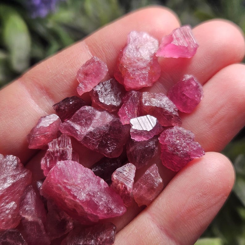 May include: A collection of small, raw, unpolished pink tourmaline crystals with irregular shapes and translucent to opaque textures. The stones exhibit varying shades of deep magenta and light pink, featuring natural facets and jagged edges.