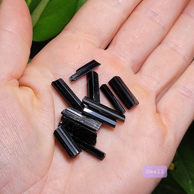 Puede incluir: Una colecci&oacute;n de peque&ntilde;os cristales de turmalina negra. Los cristales tienen varias formas, con una forma de prisma rectangular. Los cristales se sostienen en una mano, con un fondo suave y neutro.