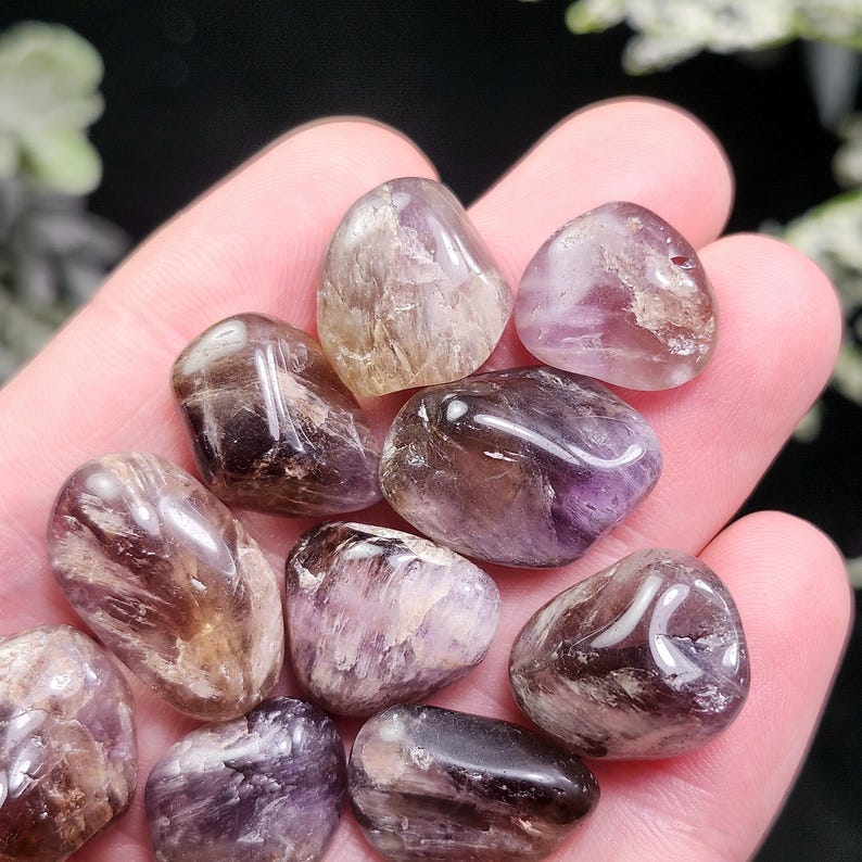 May include: A collection of polished, tumbled gemstones in shades of purple, brown, and clear. The stones are smooth and rounded, with a glossy finish. The gemstones are held in a hand, with a soft focus background.