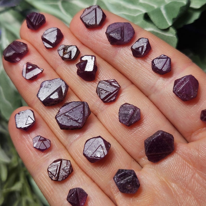 K&ouml;nnte beinhalten: Eine Sammlung von 20 kleinen, dunkelvioletten, facettierten, rohen Edelsteinen. Die Edelsteine sind auf einer Hand angeordnet, und einige der Edelsteine haben eine dreieckige Form.