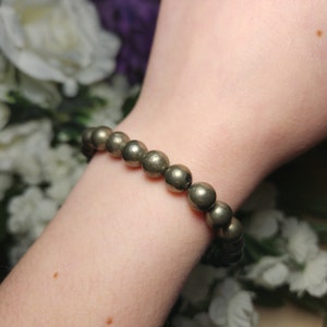 May include: A bracelet made of spherical pyrite beads, displaying a metallic, golden sheen. The beads are strung together, forming a bracelet. The bracelet is worn on a wrist, with a soft background of white flowers.
