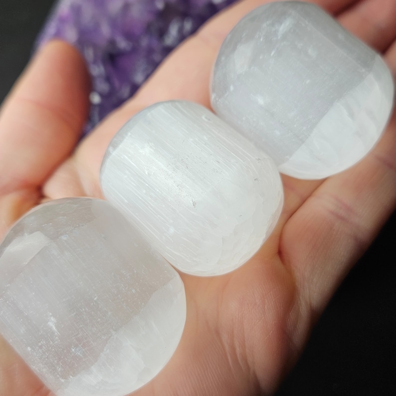 May include: Three white selenite crystal wands, also known as satin spar, are shown in a hand. The wands are smooth and have a striated texture.
