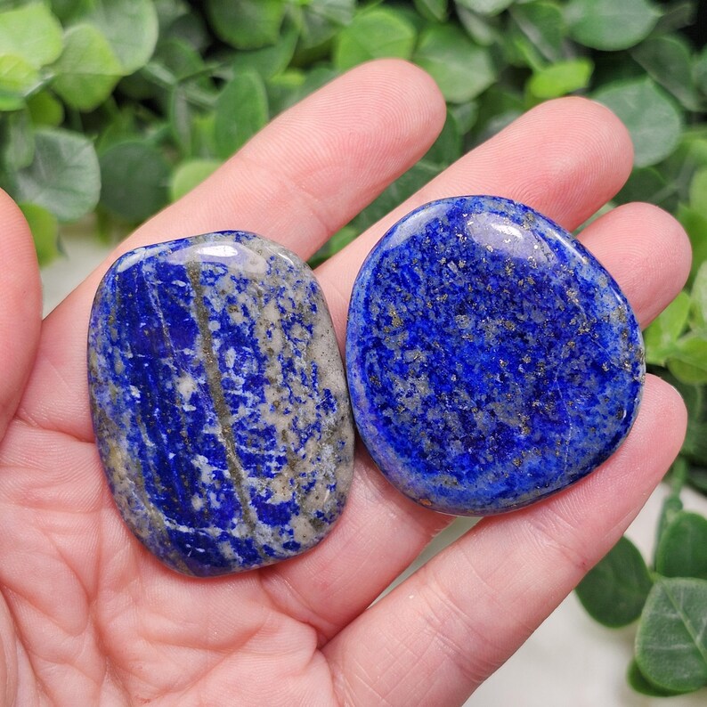 May include: Two polished lapis lazuli stones. The stones are a deep blue color with streaks of gray and flecks of gold. The stones are smooth and rounded, and they are held in a hand.