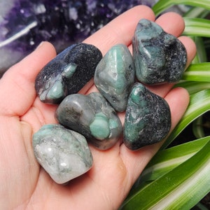 May include: A selection of emerald gemstones in shades of green and black, held in a hand. The stones are smooth and polished, with natural patterns and inclusions. The gemstones are a mix of dark and light green.