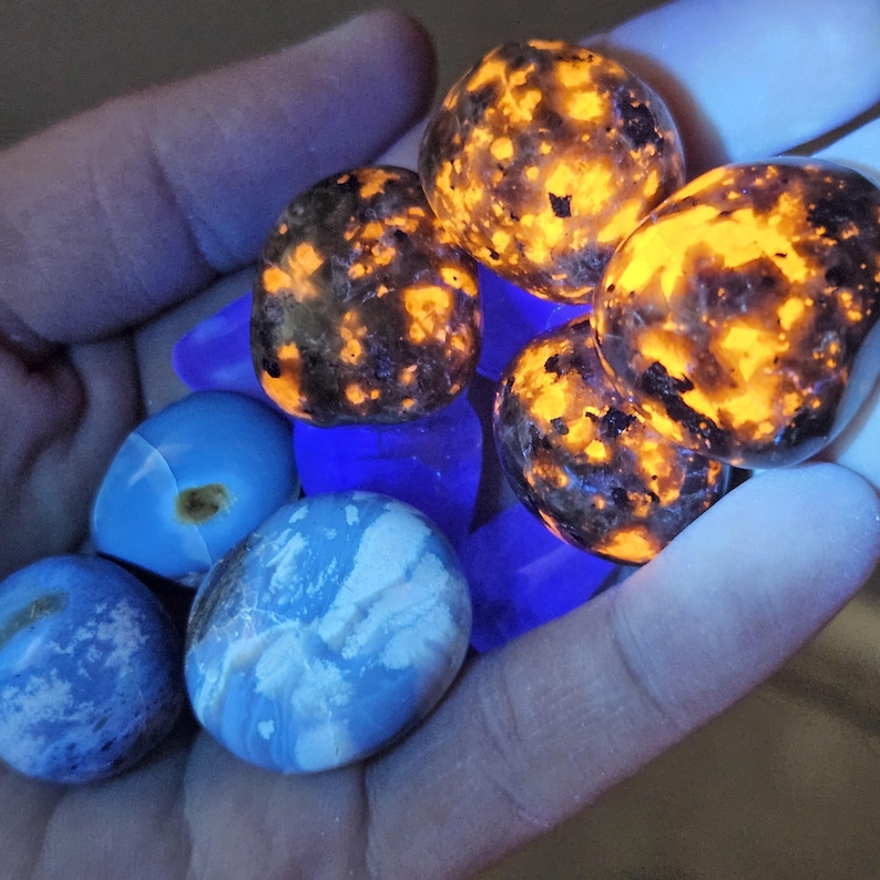 May include: A hand holding a collection of polished stones. Some stones glow with orange and yellow patterns, while others are blue and white. The stones are of various shapes and sizes, with a mix of textures.