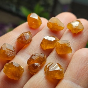 May include: A collection of raw, faceted, orange-brown gemstones. The crystals have a rough, natural texture and exhibit a warm, amber hue. The gemstones are displayed in a close-up shot, highlighting their unique shapes and reflective surfaces.