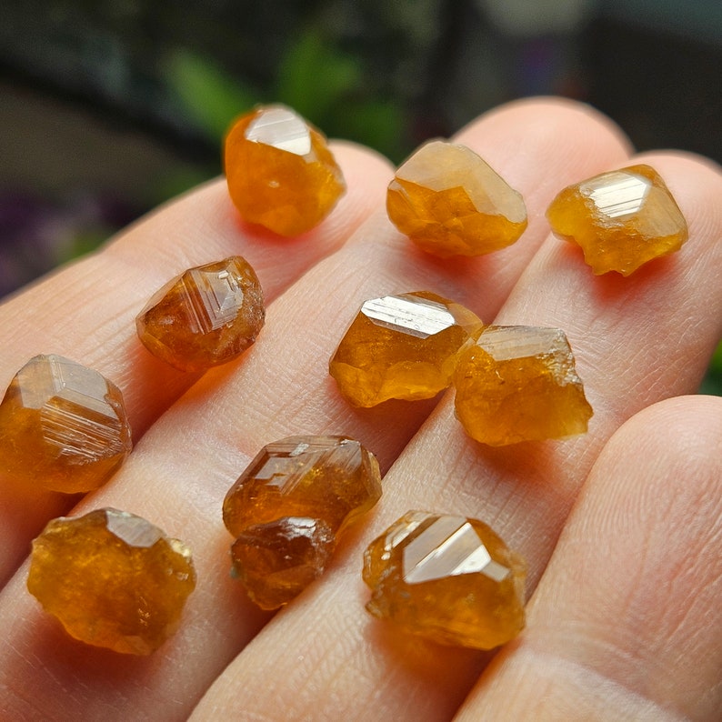May include: A collection of raw, faceted, orange-brown gemstones. The crystals have a rough, natural texture and exhibit a warm, amber hue. The gemstones are displayed in a close-up shot, highlighting their unique shapes and reflective surfaces.