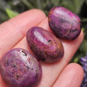 May include: Three polished, oval-shaped gemstones in varying shades of pink and purple are displayed. The stones have a smooth, reflective surface with natural patterns and inclusions. The gemstones are held in a hand, showcasing their color and texture.