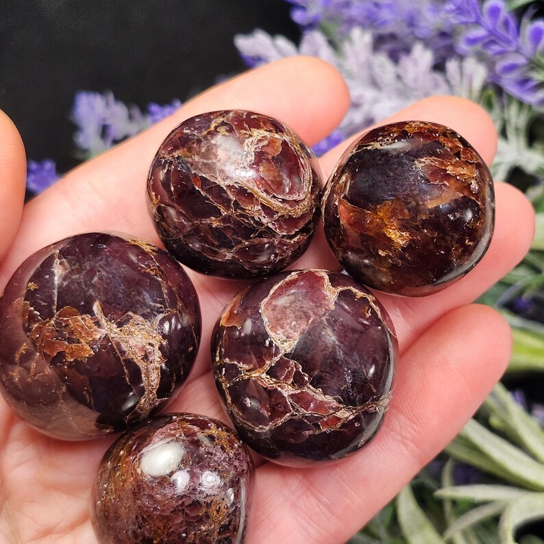 May include: Five polished, round gemstones in varying shades of deep red and brown are held in a hand. The stones have a smooth, reflective surface with natural patterns. The gemstones are a natural product.