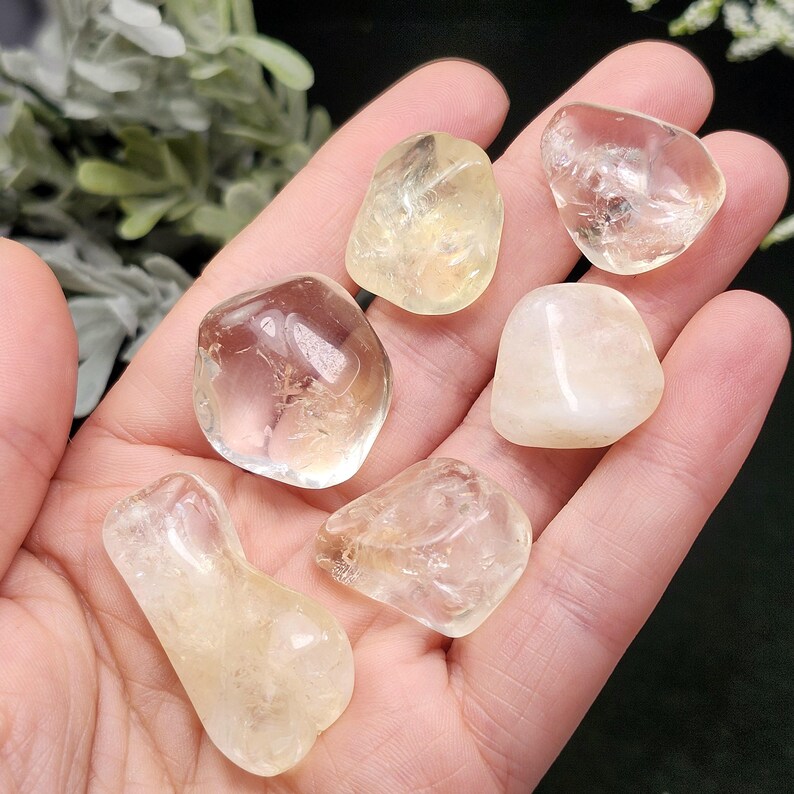 May include: A collection of tumbled citrine gemstones in various shapes and sizes. The stones range in color from pale yellow to a deeper amber hue, with a translucent quality. The gemstones are smooth and polished, reflecting light.