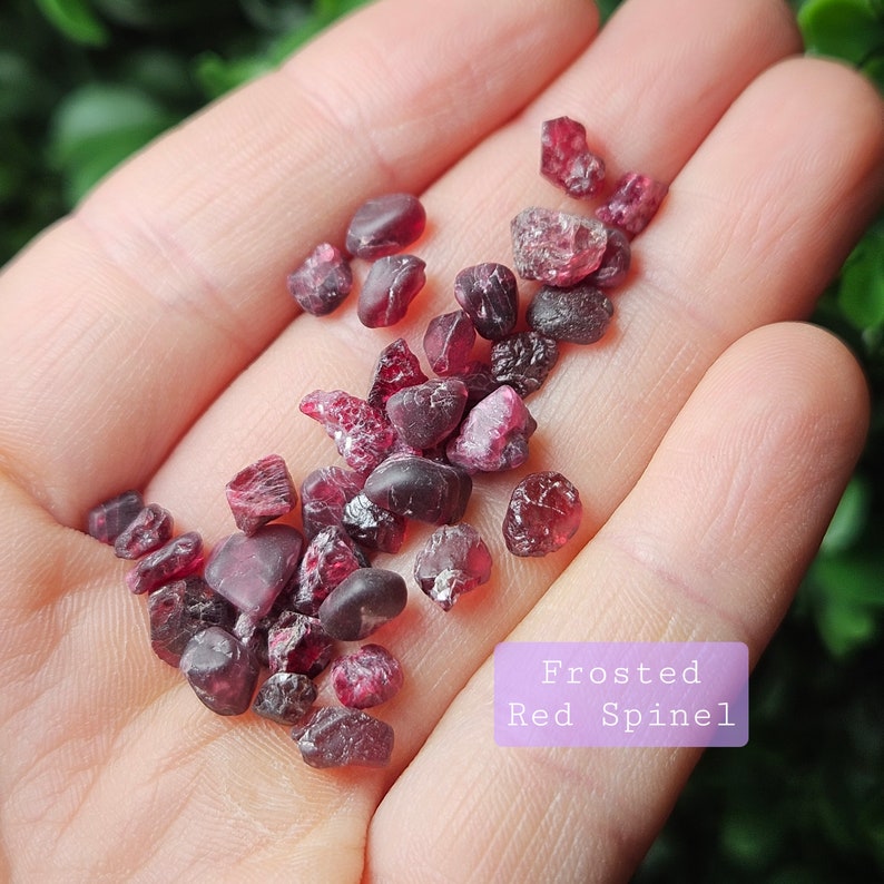May include: A collection of small, frosted red spinel crystals. The crystals are various shapes and sizes, and have a deep red colour. The crystals are displayed on a hand.