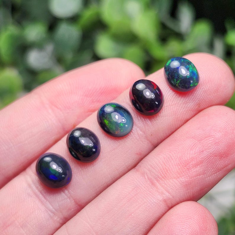 May include: Six oval-shaped black opals with iridescent flashes of green and blue. The gemstones are smooth and polished, displaying a range of dark colors. The opals are displayed on a light-toned surface, with a blurred green background.