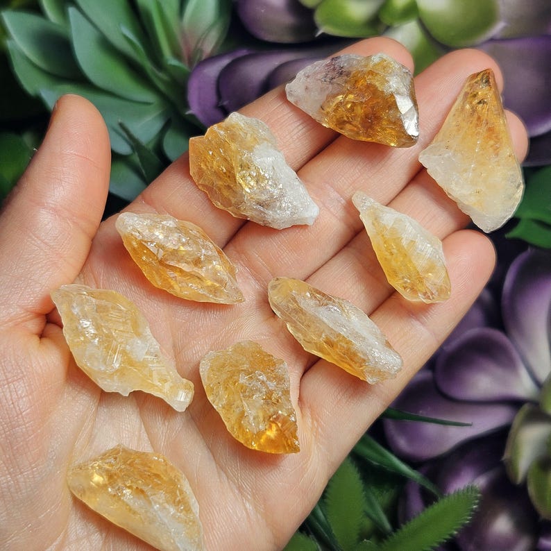 May include: A collection of nine raw citrine crystal points scattered across an open palm. Each piece features a translucent, golden-yellow hue with jagged, natural edges and varying geometric shapes, showcasing the raw, unpolished texture of quartz.