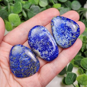 May include: Three polished lapis lazuli stones in varying shapes are displayed. The stones are a deep blue color with flecks of gold and white. The stones are smooth and have a glossy finish, perfect for jewelry or display.