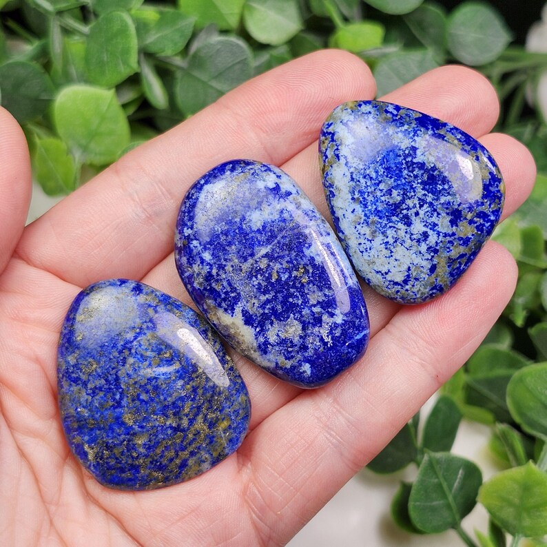 May include: Three polished lapis lazuli stones in varying shapes are displayed. The stones are a deep blue color with flecks of gold and white. The stones are smooth and have a glossy finish, perfect for jewelry or display.