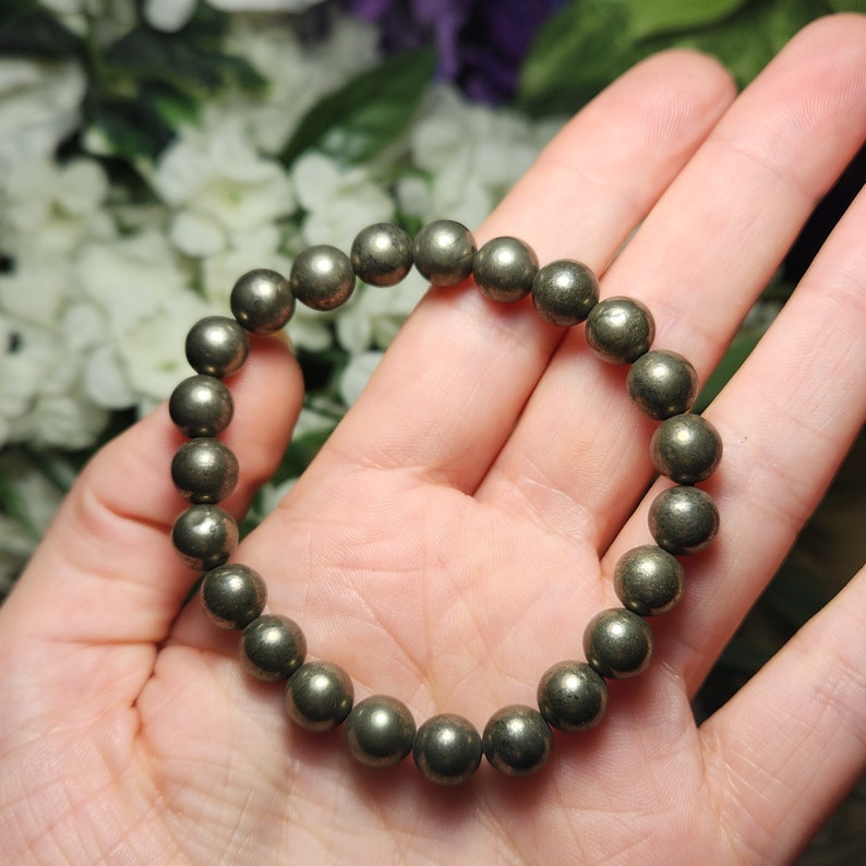 May include: A bracelet composed of spherical pyrite beads, in a muted gold hue. The beads are strung together, forming a circular bracelet. The jewellery is displayed on a hand, with a blurred floral backdrop.