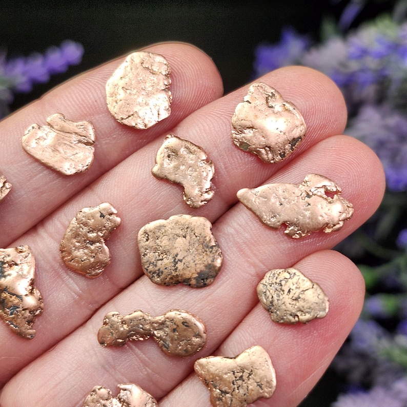 May include: A collection of small, irregularly shaped copper nuggets. The nuggets have a warm, reddish-brown color and a rough, textured surface. They are displayed on a hand, with a blurred background of purple flowers.